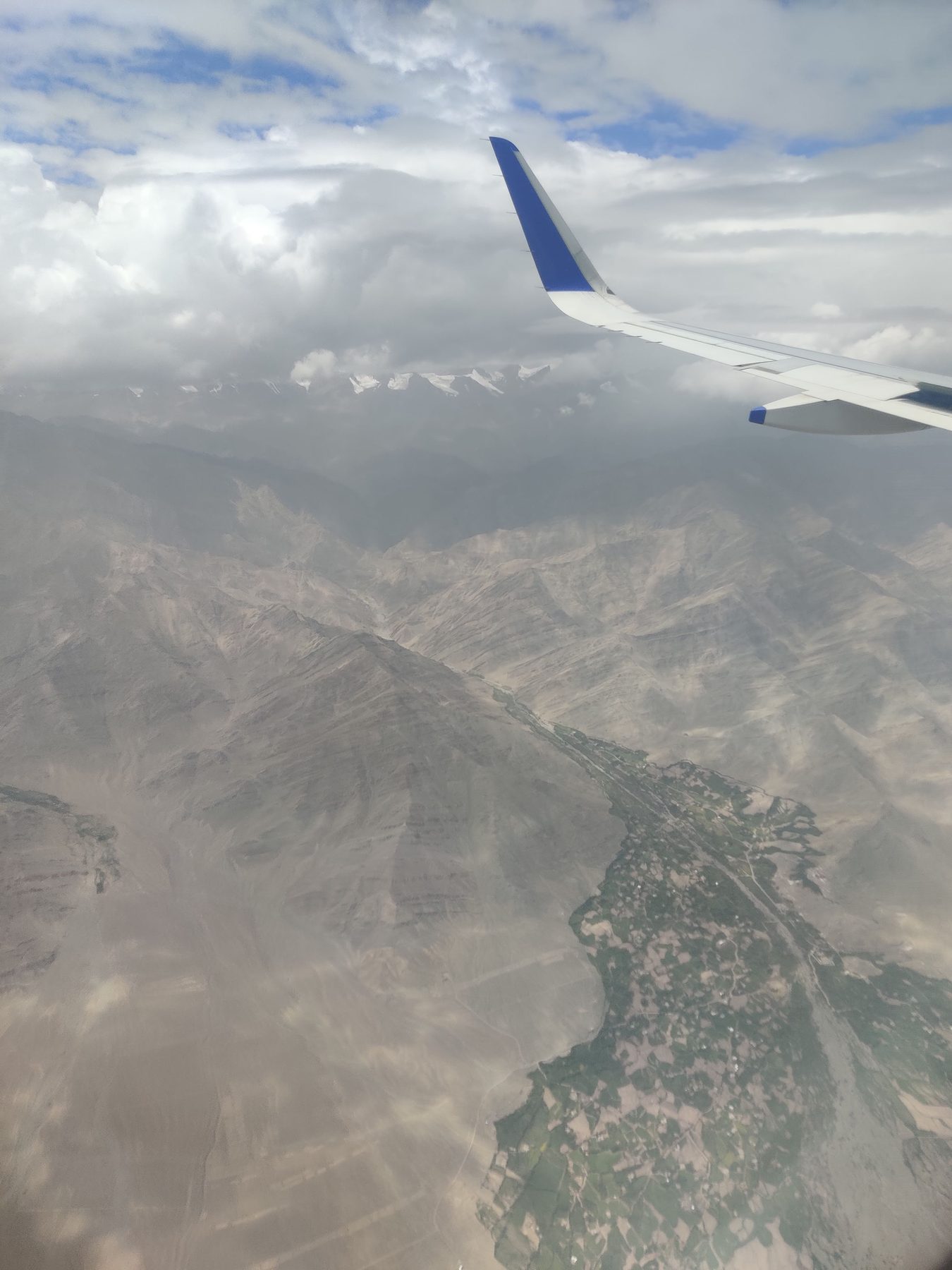 View from a plane window over bare brown mountains and a thin green river valley, snow peaks on the horizon