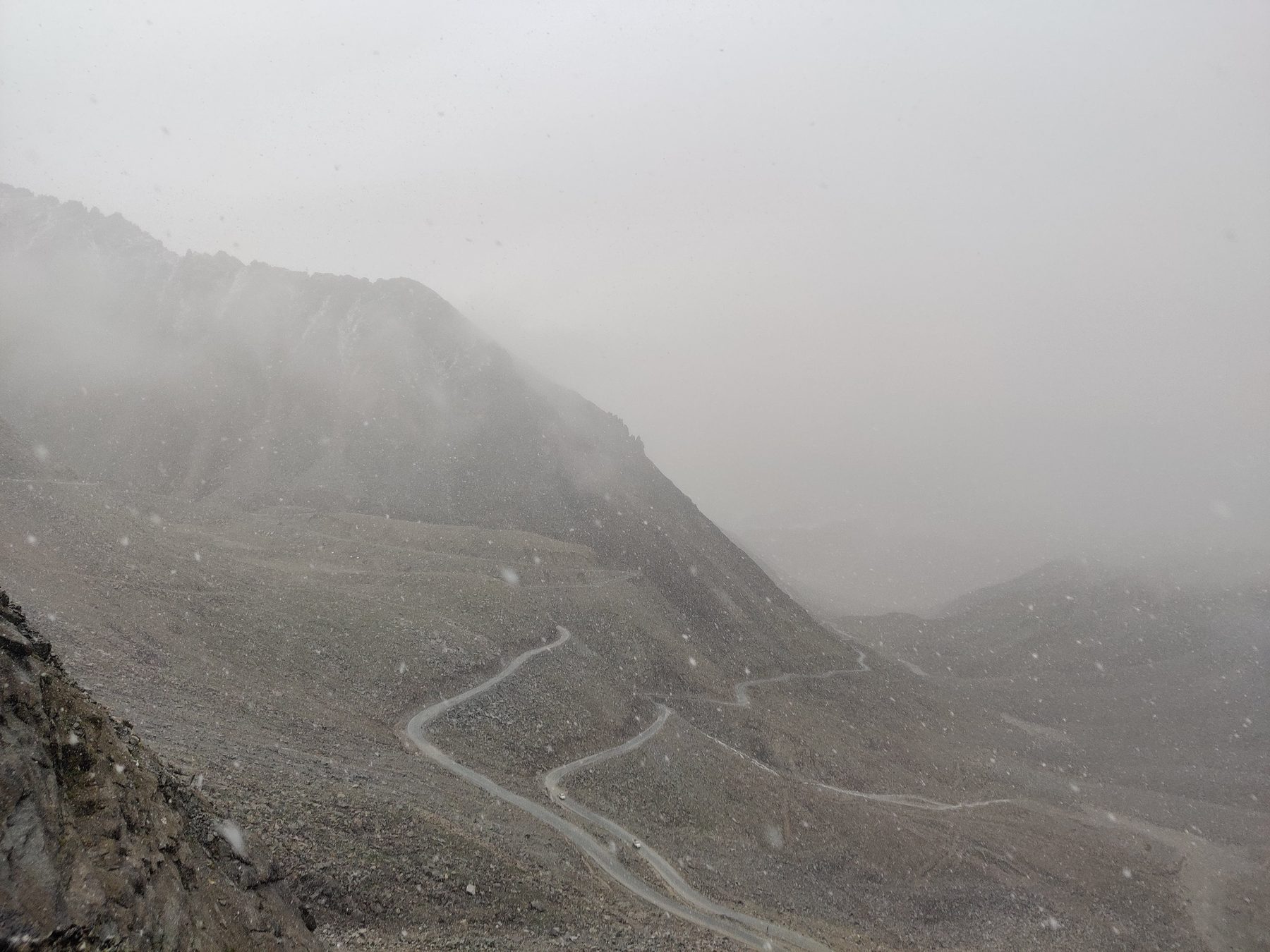 A woman in a beige coat among prayer flags at Khardung La, arms stretched wide, face tilted up to the falling snow with eyes closed