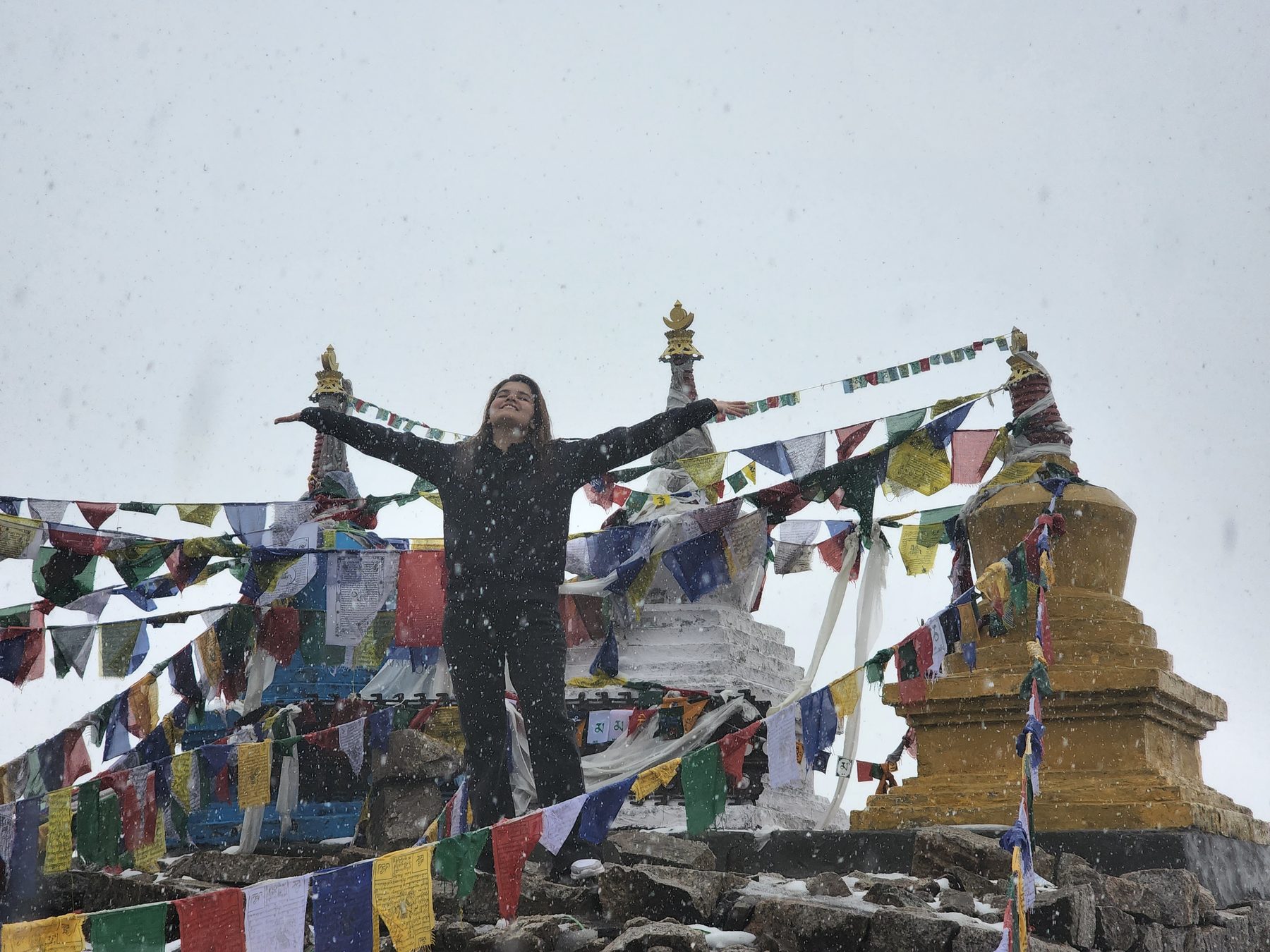 A second woman in black, same place, arms outstretched, face lifted to the falling snow, smiling