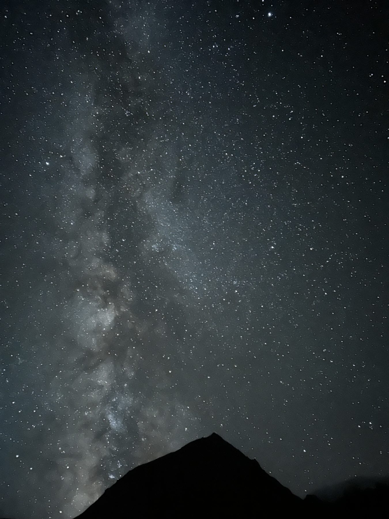 The Milky Way stretching across a black sky crowded with stars, a dark mountain silhouette at the bottom of the frame